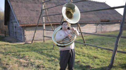 The guy with the tuba makes sharp movements to the right and left down and up.Funny musician movement in plaid pants and a linen shirt . Against the landscape of the village.