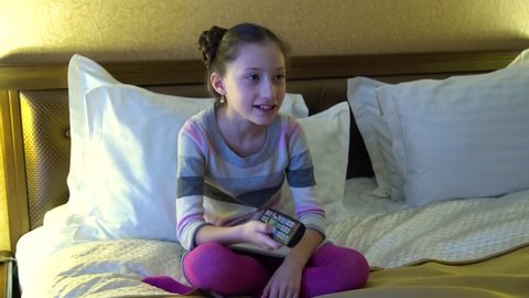 Beautiful little girl sitting on the bed in the hotel and watching TV. In the hands of a girl holding a remote control from the TV.