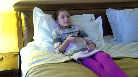 Beautiful little girl sitting on the bed in the hotel and watching TV. In the hands of a girl holding a remote control from the TV.