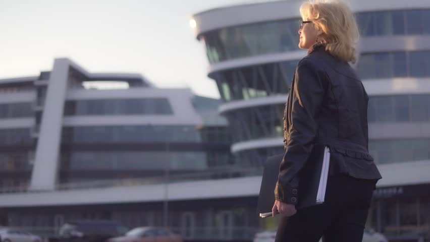 A woman walks down the street towards the sun and holds a folder for documents in her hand. Slow-motion