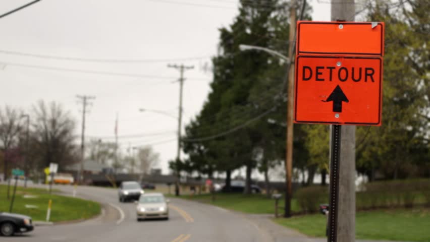 A orange construction detour sign with arrow pointing straight in generic overcasy residential road