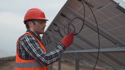 The worker in uniform and red hardhat talks on a radio and begins mounting of a photovoltaic panel on solar farm