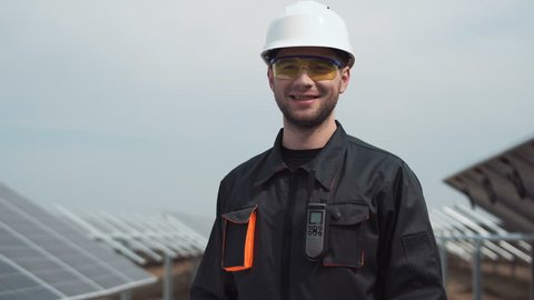 The employee of power plant in a uniform and white hardhat, standing between rows of photovoltaic panels looks in the camera and smiles