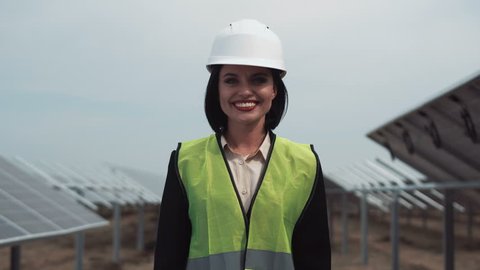The woman engineer of power plant in a uniform and white hardhat, standing between rows of photovoltaic panels looks in the camera and smiles