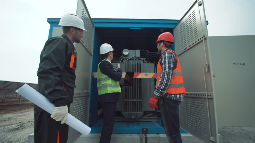 The group of electricians and engineers stand near the transformer of the electric power and discuss blueptints using the computer