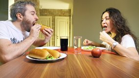 Young caucasian man and woman eating hotdogs and lettuce at home. Junk food versus healthy eating concept. 4K video