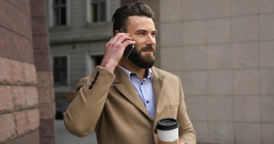 Stylish young attractive man with a beard speaks on the phone and drinking coffee at the business center on a sunny day