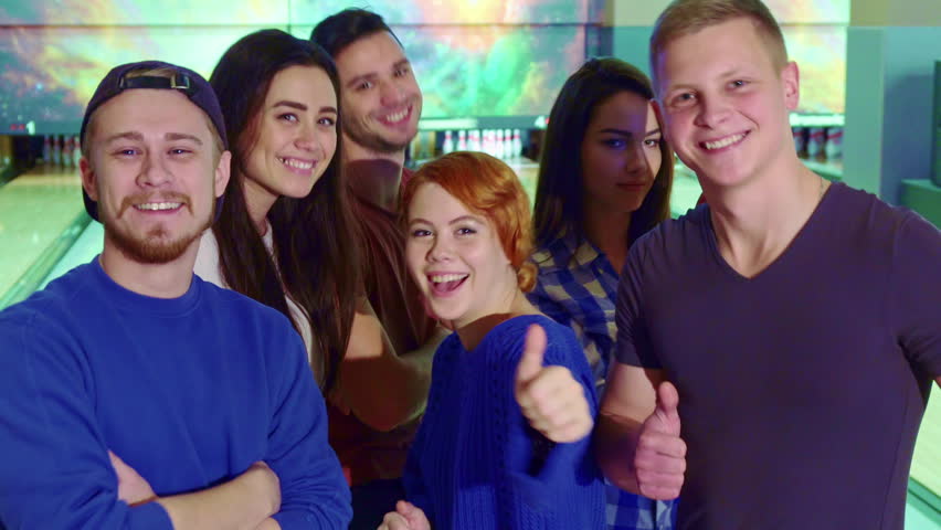 Friends having fun at the bowling. Young people dancing against background of lanes. Close up of attractive teenagers showing their thumbs up