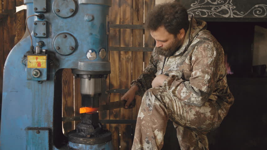 Bearded young man blacksmith using hydraulic press for steel arms manufacture at smithy workshop
