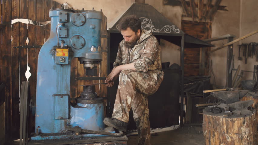 Bearded young man blacksmith using hydraulic press for steel arms manufacture at smithy workshop
