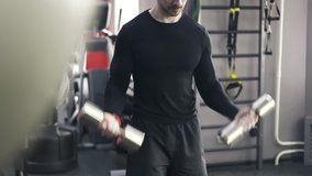Bearded young man wearing black sportswear doing dumbbell lifts in a gym. Tilt up real time medium shot