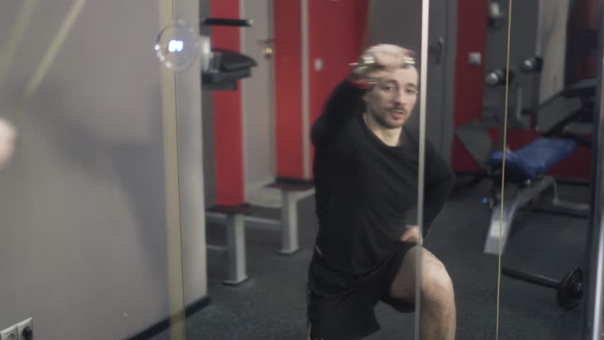 Front view of a bearded young man in black sportswear is lifting weights with one hand in a gym. Handheld real time medium shot.