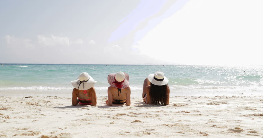 Back Rear View Of Three Girls Lying On Beach Raising Legs, Enjoy Sun Tan, Woman Tourists On Summer Holiday Slow Motion 60