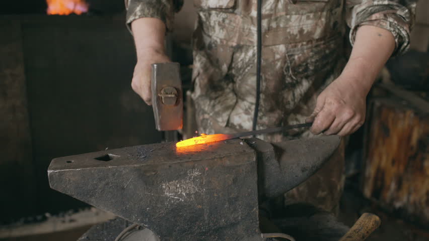 Tilt up young man blacksmith manually forging hot metal knife on anvil in smithy with spark fireworks