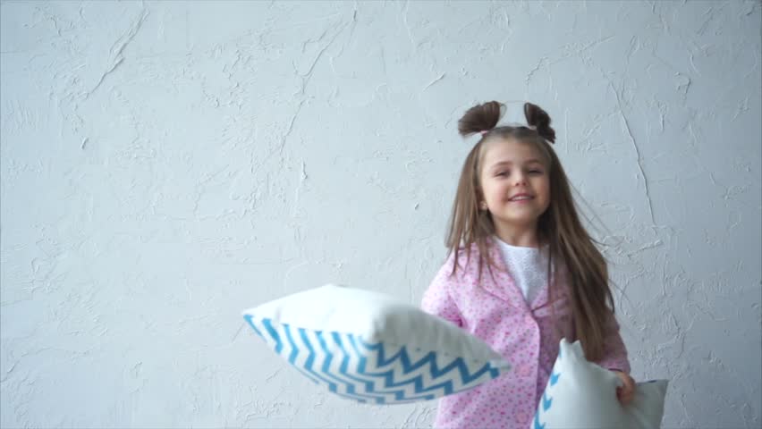 A nice and beautiful girl with funny tails on her head is standing on the bed and playing with pillows, throwing them up, and then a beautiful baby is spinning around.
