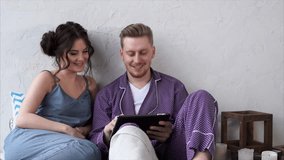 A couple in love who are dressed in pajamas sit on a bed in a bedroom and choose a hotel on a tablet for a joint holiday. A woman and a man plan a romantic trip.