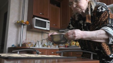 Grandmother puts a metal plate on the table, takes a spoon, sits down to eat