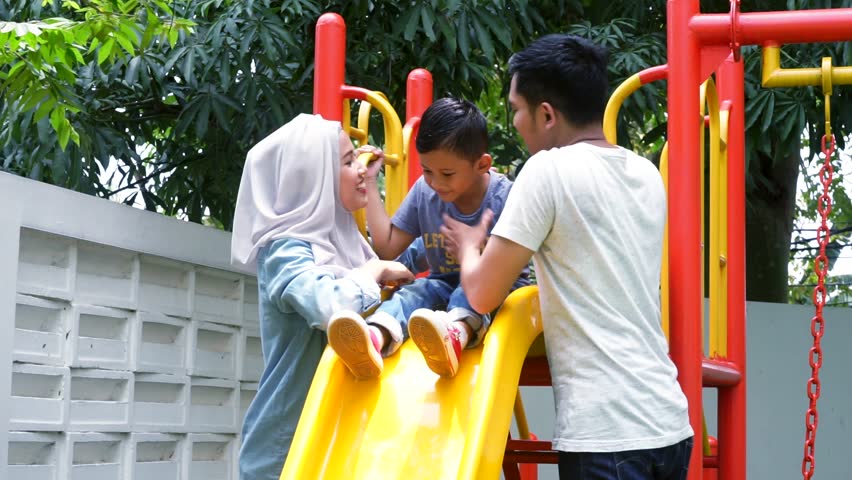 Joyful little boy playing on slide and kissed by his parents, shot outdoors