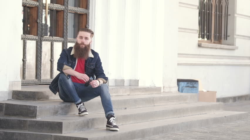 Handsome young stylish hipster man with long beard in grey shirt standing outdoor. Denim style