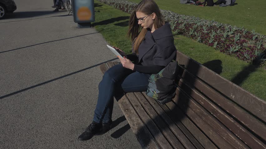 Young brunette woman with eyeglasses reading a book outdoor. Sitting on the bench in the park. Spring time.
