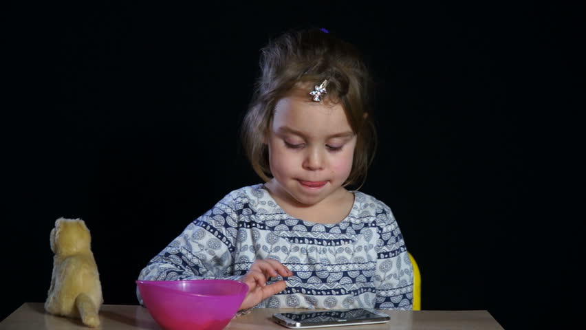 Sleepy girl of European appearance 3-4 years, refuses to sleep. A child is sitting at a table, playing on a smartphone, eating an apple. Dark room, black background.