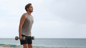 Fitness man lifting dumbbells on beach doing Front Dumbbell Raise i.e. Alternating Front Raise workout for shoulders. Exercising male fitness model working out on beach. SLOW MOTION 59.94 FPS.