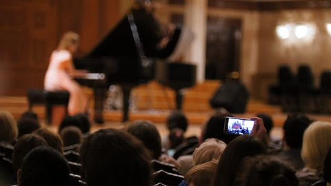 Audience in concert hall during performing piano girl- people shooting performance on smartphone, music opera