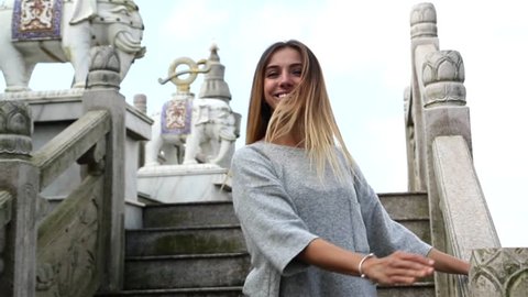 Beautiful long hair blond girl is running up the stairs, turning around, waving hair and smiling happily. White stone stairs and elephants statues in background 
