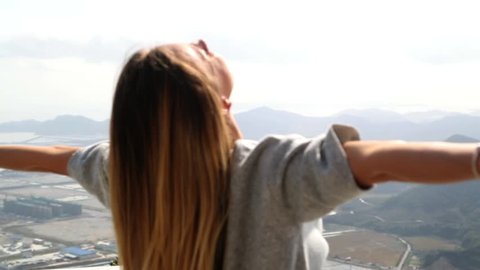 Girl with blond long hair, raising her hands up, feeling happy and free, enjoying the view, mountains sea and fields in background 