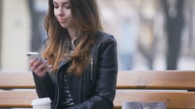 Smiling young woman with cup of coffee while using smartphone on the bench in the street