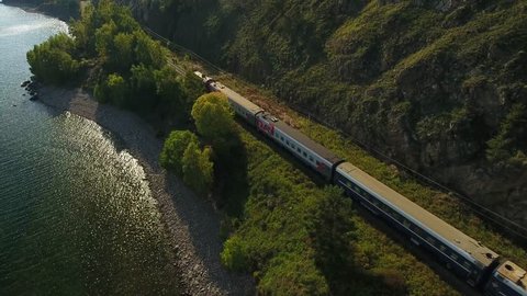 Lake Baikal oldest. Railways Russia Siberia. listvyanka. Passenger train rides near the water behind trees. Mountains. Summer sun shines blue water. Unique aerial video view from above train 4k