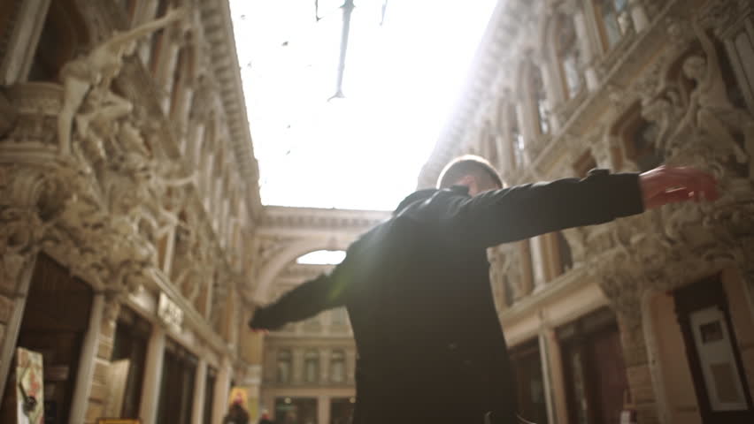 Young handsome Caucasian male in street outerwear smiling dancing in circles in beautiful passageway in Odessa. View from below with sun flare slowmotion