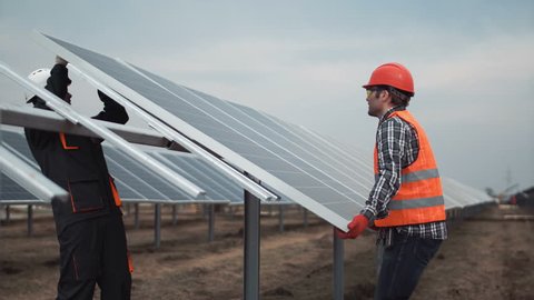 Two workers in a uniform and hardhat install photovoltaic panels on a metal basis on a solar farm