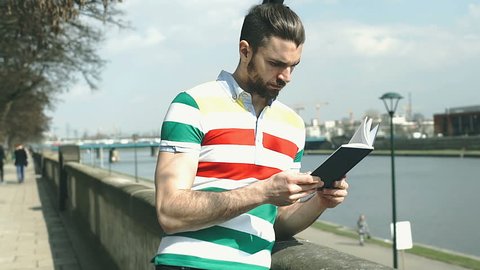 Handsome man reading book next to the river and smiling to the camera, steadycam shot
