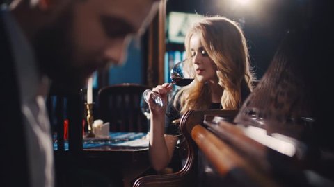 Gorgeous young woman in a classic black dress drinking red wine and seductively looking at the young bearded musician playing the vintage piano. Romantic atmosphere, perfect date. Couple goals.