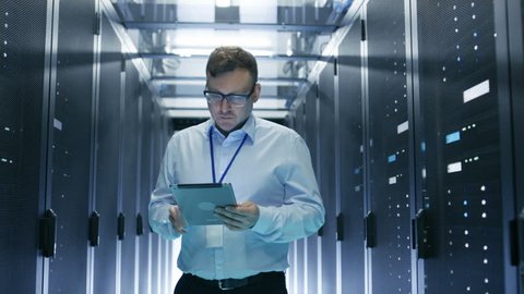 IT Technician Walks Through Rows of Server Racks in Data Center. Simultaneously He Works on a Tablet Computer. Shot on RED EPIC-W 8K Helium Cinema Camera.
