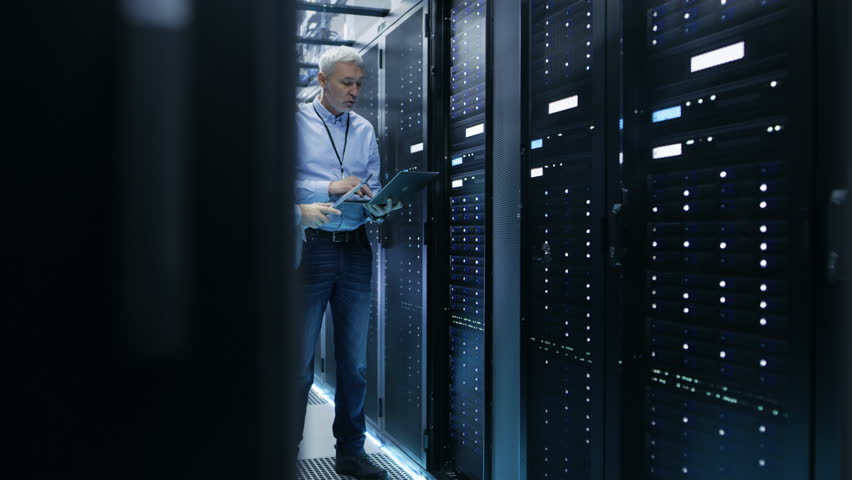 Two IT Engineers Standing in Working Data Center. They Use Laptop and Tablet Computer while Standing Beside Open Server Rack Cabinet. Shot on RED EPIC-W 8K Helium Cinema Camera. Royalty-Free Stock Footage #26008067