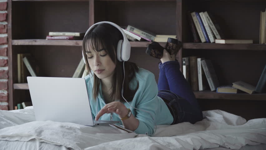 Young brunette woman in headphones is laying on a bed with a laptop and texting, book shelves behind