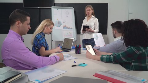 Young successful business team working in the office. Woman standing near flipchart using digital tablet tell about new products or services. Five professional workers in boardroom.