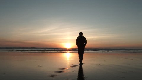 Silhouetted man casually walks out on low tide beach to enjoy the peaceful sunset over the Pacific Ocean.