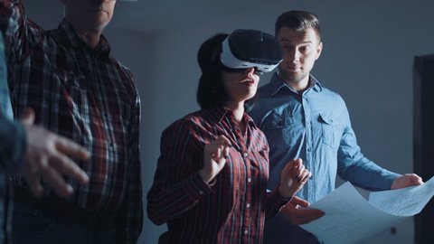 The people with blueprint and virtual reality headsets on a construction site. The woman shows to group of architects and engineers the project of future interior of the room in the 3D simulator