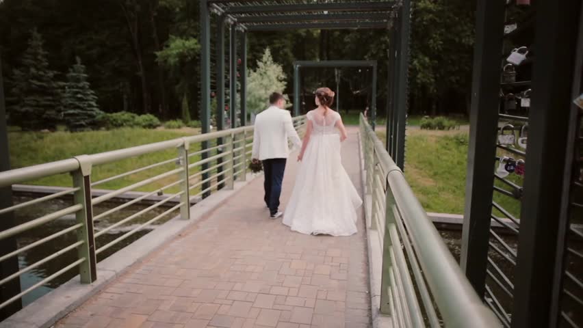 Back view of lovers walking on a bridge in a park. Bride and groom have good time on their wedding day. | Shutterstock HD Video #26034230