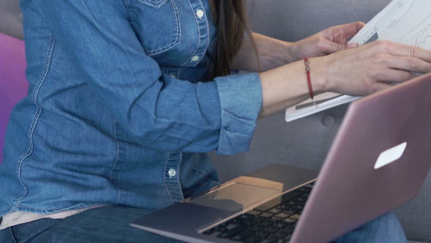Young woman working with laptop and documents sitting on sofa at home
