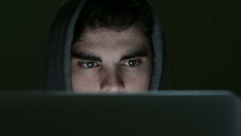 Camera tracks across face of suspicious looking young man wearing hooded top working on laptop at night