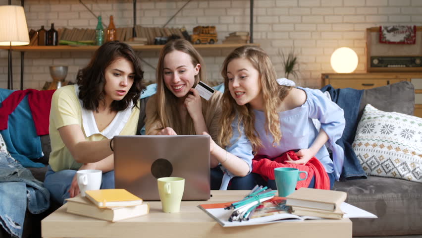 three active happy adult girlfriends look for and choose new clothes in online shop laughing and make payment online by credit card using laptop computer and internet while sitting at home