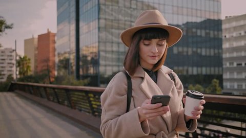 Trendy student in fedora hat, round glasses and hipster backpack scrolls through her news feed on smartphone on way to university college to stay up to date with events and social media