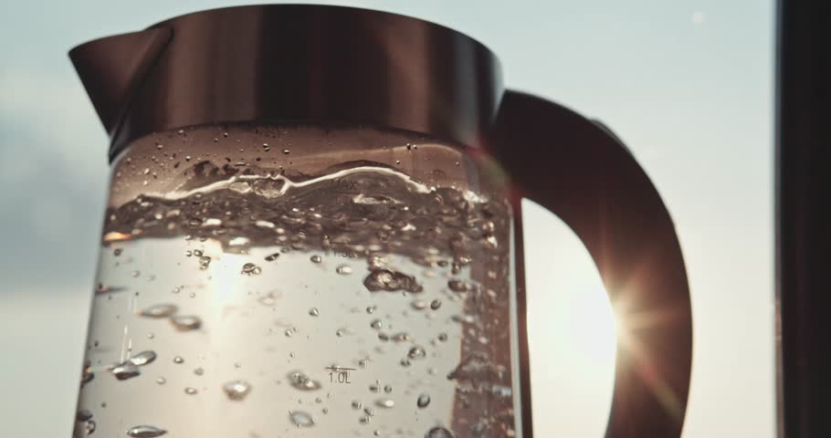 SLOW MOTION: Steaming glass electrical tea kettle boiling in the morning, close up. Steam comes out of a kettle in gentle light of sunrise. Cozy morning at home kitchen. 120 fps 4K DCi.