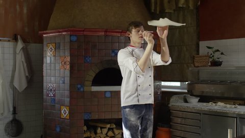 Young guy in chef uniform spinning and tossing pizza dough in restaurant kitchen in slow motion
