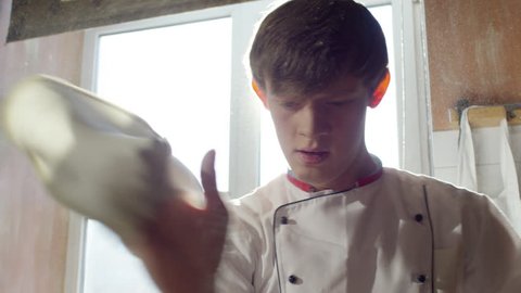 Closeup of young pizzaiolo spinning pizza dough on fingertip and throwing it up in sunny restaurant kitchen. Slow motion footage