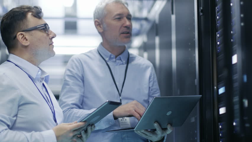 Two IT Technicians Standing in Working Data Center. They Use Laptop and Tablet Computer while Standing Beside Open Server Rack Cabinet.  Shot on RED EPIC-W 8K Helium Cinema Camera. Royalty-Free Stock Footage #26069843
